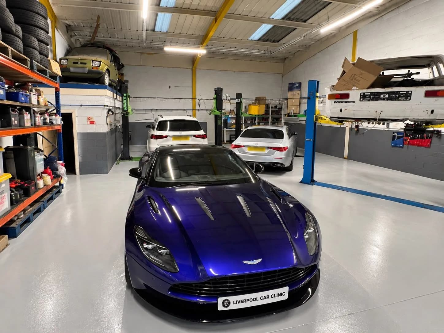 A blue Aston Martin carrying a Liverpool Car Clinic trade plate on the workshop floor, with two Mercedes behind it and classic cars raised on the ramps.