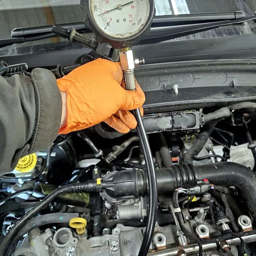 A technician in an orange nitrile glove holding a compression test gauge against an engine bay.