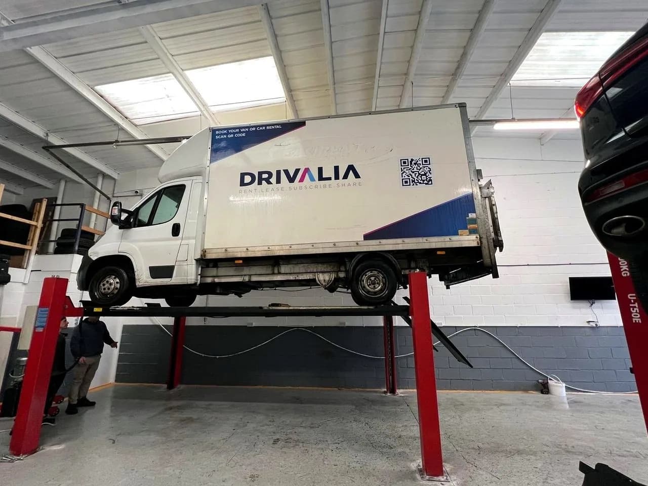 A Drivalia Luton van raised on the four-post ramp inside the Liverpool Car Clinic workshop, with two technicians working underneath.